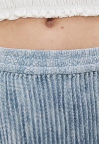 Lyseblå plisséskjørt med elastisk midjebånd. Stoffet har en subtil tekstur og mønster som komplementerer en hvit volang croptopp.