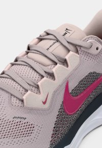 Gros plan sur une chaussure de course Nike rose clair avec des lacets violets, un logo swoosh rose foncé et une tige en tissu en maille respirante.