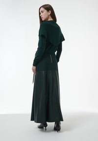 Donkergroen gebreid sweater met een gedrapeerde achterkant, gecombineerd met een lange zwarte leren rok met een gouden ritsdetail. Zwarte hakken maken de outfit af.