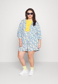 Conjunto de camisa e calções com padrão floral azul, com uma t-shirt amarela por baixo; tênis brancos, meias amarelas e óculos de sol pretos.