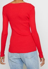 Chemise rouge à manches longues côtelée avec un col large, coupe ajustée et tissu texturé. Associée à un jean taille haute bleu clair.