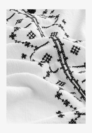 Fehér szövet bonyolult fekete hímzéssel, amely geometrikus mintákat és texturált varrást tartalmaz, létrehozva egy rétegezett megjelenést.