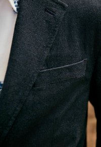 Dunkel strukturiertes Blazer mit einem Reverskragen, ausgestattet mit einer Brusttasche und einer dezenten Stoffwebung. Sichtbares Innenfutter in heller Farbe.