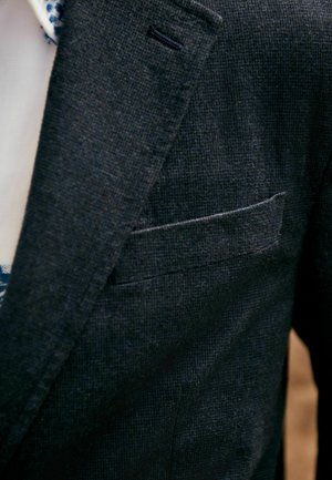 Tamna strukturirana blazerica s ovratnikom, koja ima džep na prsima i suptilno tkanje tkanine. Vidljiva unutrašnja podstava svijetle boje.