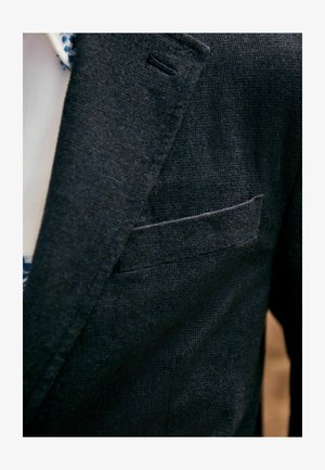 Tamna strukturirana blazerica s ovratnikom, koja ima džep na prsima i suptilno tkanje tkanine. Vidljiva unutrašnja podstava svijetle boje.