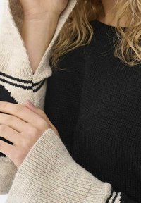 Pull en maille avec un corps noir et une capuche ainsi que des manches crème claires et texturées. Les manches comportent des accents à rayures noires près des poignets.