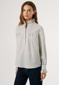 Srajca z dolgimi rokavi v svetlo sivi barvi s karirastim vzorcem. Ima stoječ ovratnik, delno zapiranje spredaj in naborke na prsim in manšetah.