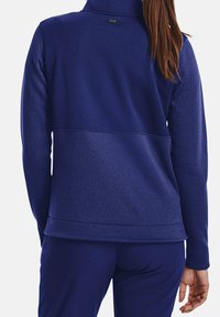 Modrá fleecová bunda s vysokým límcem, textured tkaninou a kontrastní hladkým panelem v pase; na zadní straně je malá logová visačka.