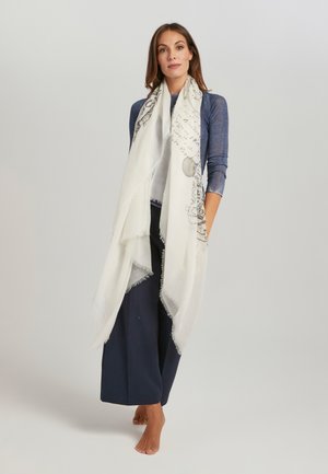 Witte sjaal met een ingewikkeld grijs patroon. Gemaakt van lichtgewicht stof met rafelige randen, elegant vallend over een donker outfit.