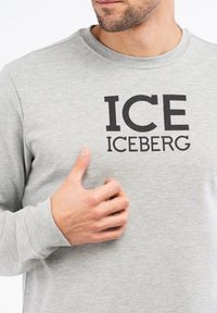 Siva majica dugih rukava od mekog materijala s velikim crnim natpisom "ICE ICEBERG" preko grudi.