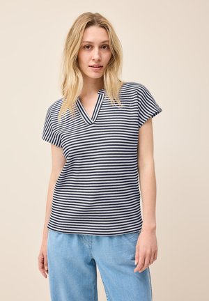 Haut à rayures marines et blanches à manches courtes avec un col en V et un tissage texturé ; porté avec un jean léger et ample devant un fond neutre.