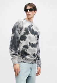 Sweatshirt cinza com efeito tie-dye e padrões pretos, com decote de gola redonda e punhos canelados, combinado com jeans azul claro e óculos de sol escuros.