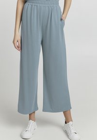 Donna che indossa pantaloni larghi e cropped di colore azzurro chiaro con cintura elastica e sneakers bianche, in piedi con una mano in tasca.