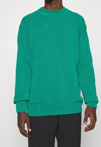 Turkos stickad tröja, rundad hals, ribbade muddar och fåll, texturerat mönster, avslappnad passform, bärs med mörka byxor.