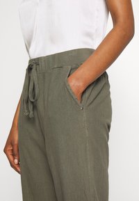 Pantalon vert olive, léger, coupe décontractée avec taille élastique, cordon de serrage et poches latérales. Tissu texturé avec une finition douce.