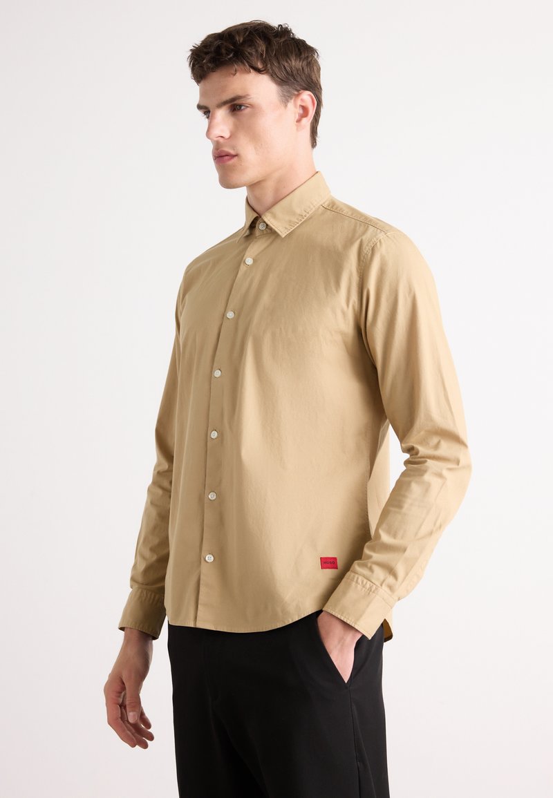 Beige button-up shirt made of smooth fabric, featuring a classic collar, long sleeves, and a red logo tag at the bottom left.