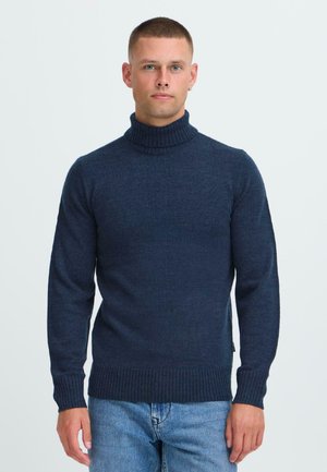 Mies, joka seisoo tummansinisessä ribbed-mallin kauluspuserossa ja vaaleansinisissä farkuissa, taustanaan yksinkertainen valkoinen tausta.