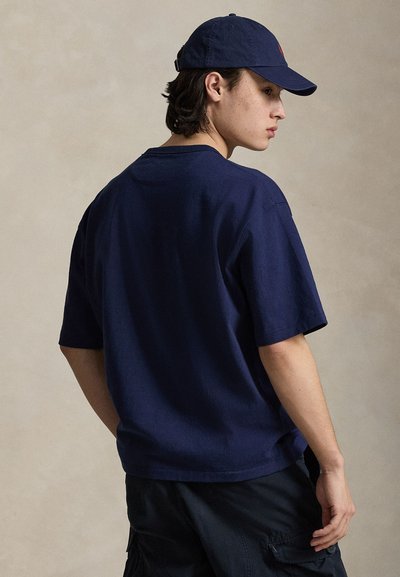 Νεαρός άνδρας που φοράει μπλε ναυτικό μπλουζάκι, ταιριαστό καπέλο μπέιζμπολ και σκούρα σορτς cargo, κοιτώντας πάνω από τον ώμο του μπροστά σε απλό φόντο.