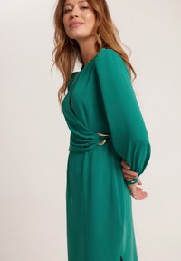 Zielona sukienka z długimi rękawami, zebranym detalem w talii i złotymi akcentami. Gładki, płynny materiał; widoczny rozcięcie po boku.