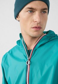 Giacca leggera color verde smeraldo con cappuccio, caratterizzata da una cerniera bianca e dettagli rossi. Il modello indossa un berretto a coste blu scuro. Vista ravvicinata.