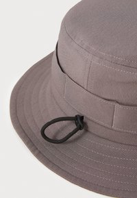 Grå bucket hat lavet af tekstureret stof, med en bred skygge, syede detaljer og et justerbart sort snor på siden.