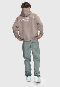 Mann trägt einen taupefarbenen Hoodie mit dem Text "Dropsize Studios Ltd. Erfolg ist ein Straßenspiel" auf dem Rücken, hellblaue Jeans und weiße Sneakers und steht mit dem Rücken zur Kamera.