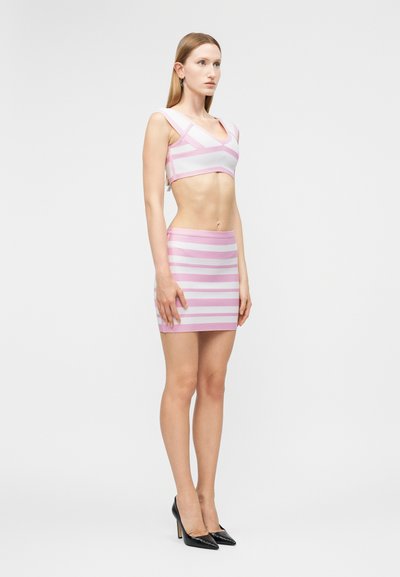 Γυναίκα που στέκεται φορώντας μια ροζ και λευκή ριγέ κοντή μπλούζα και ταιριαστό μίνι φούστα με μαύρα τακούνια, στραμμένη προς τα αριστερά σε απλό φόντο.