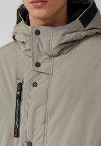 Hellgraue isolierte Jacke mit Kapuze, schwarzen Akzenten, Druckknöpfen und einem Frontreißverschluss mit sichtbaren Nahtdetails.