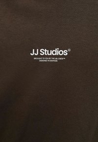 Braunes T-Shirt aus weichem Stoff mit weißem Text: "JJ Studios" und zusätzlicher Beschriftung, vor einem einfarbigen Hintergrund.