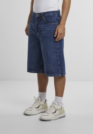 Dunkelblaue Jeansshorts mit lockerem Sitz, Fünf-Taschen-Design und leichter Waschung. Kombiniert mit weißen und hellgelben Sport Sneakern.