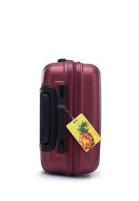 Maleta rígida de color rojo metálico, con una superficie texturizada, cremalleras dobles y un asa negra, con una etiqueta de equipaje temática de piña adjunta.