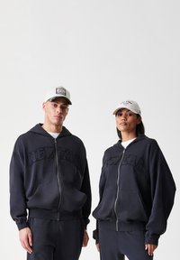 Felpe con cappuccio blu scuro a zip presentano la scritta in rilievo "NEWERA", abbinate a pantaloni della tuta. Entrambi i modelli indossano cappelli bianchi e beige.