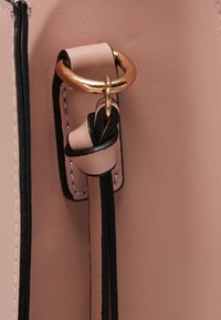 Detail růžové kožené tašky s zlatým kovovým kroužkem a tmavými popruhovými akcenty, s kontrastním prošíváním podél okrajů.