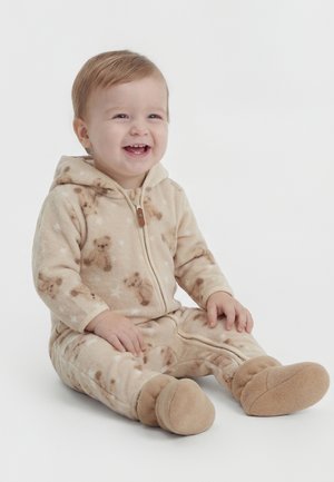 Smiling toddler sitting wearing beige fuzzy hooded onesie with teddy bear and star patterns and matching soft booties.