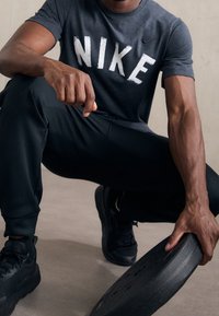 Mustat Nike-tunikat, joissa on valkoista kirjontaa, mustat urheiluhousut ja mustat lenkkarit. Henkilö pitää mustaa painolautaa, esitellen lihaksikkaita käsiään.