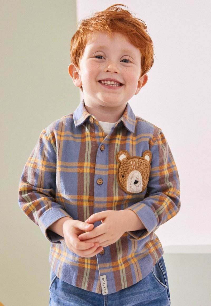 Lächelnder rothaariger Junge trägt ein blau-orange kariertes Hemd mit einem Teddybär-Aufnäher auf der Brust und blaue Jeans, steht drinnen.