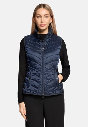 Kvinde iført en marineblå quiltet ærmeløs vest over en sort langærmet sweater, stående med den ene hånd i vestens lomme mod en ensfarvet baggrund.