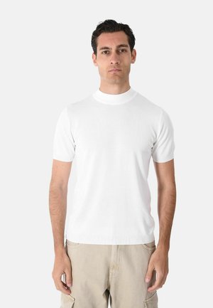 Uomo con capelli scuri in piedi, indossa una maglietta bianca semplice a maniche corte con collo alto e pantaloni cargo beige su uno sfondo chiaro uniforme.