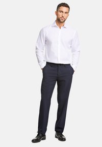 Weißes Hemd mit Knöpfen und langen Ärmeln, kombiniert mit dunkelblauen Hosen und schwarzen Anzugschuhen. Einfaches Design, taillierte Passform und keine sichtbaren Muster.