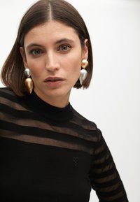 Čierny top s dlhými rukávmi, ktorý má priesvitné pásy, vysoký golier a malé vyšívané srdiečko na hrudi. Súčasťou sú výrazné náušnice.