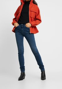 Chaqueta roja de cremallera con dos bolsillos en el pecho, combinada con jeans ajustados azul oscuro y botas negras de tobillo. Atuendo moderno y casual.