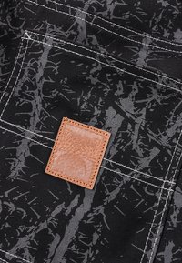 Μαύρο ύφασμα με υφή γκρι αφηρημένου σχεδίου, διακοσμημένο με λευκή ραφή και καφέ δερμάτινο ταμπελάκι με ανάγλυφο σχέδιο.