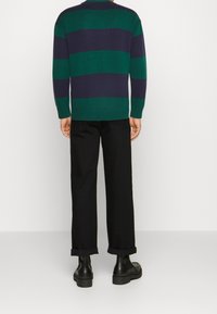 Pull en maille avec des rayures horizontales vertes et bleu marine, poignets et ourlet côtelés. Associé à un pantalon noir retroussé et des bottes noires épaisses.