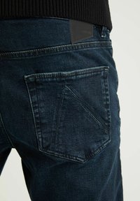 Mörka denimjeans med en standard bakfickdesign, en texturerad yta och en svart läderlogga nära midjan.