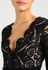 Model předvádí detailní pohled na černé krajkové šaty se složitými květinovými vzory a hlubokým V-výstřihem, který ukazuje elegantní detaily a textury.