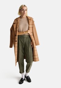 Cappotto di lana tan con pattern a scacchi, maglione beige, pantaloni in velluto a coste verde oliva, mocassini neri e calzini bianchi, in piedi su uno sfondo neutro.