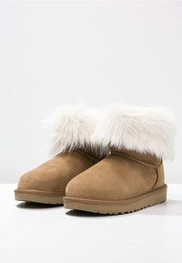 Bottines en daim camel avec un revers en fausse fourrure blanche, semelle plate et coutures le long des bords. Adaptées pour temps froid.