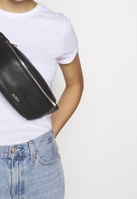Persona che indossa jeans azzurri chiari e una maglietta bianca con una borsa a tracolla in pelle nera etichettata "HUGO" sul petto.