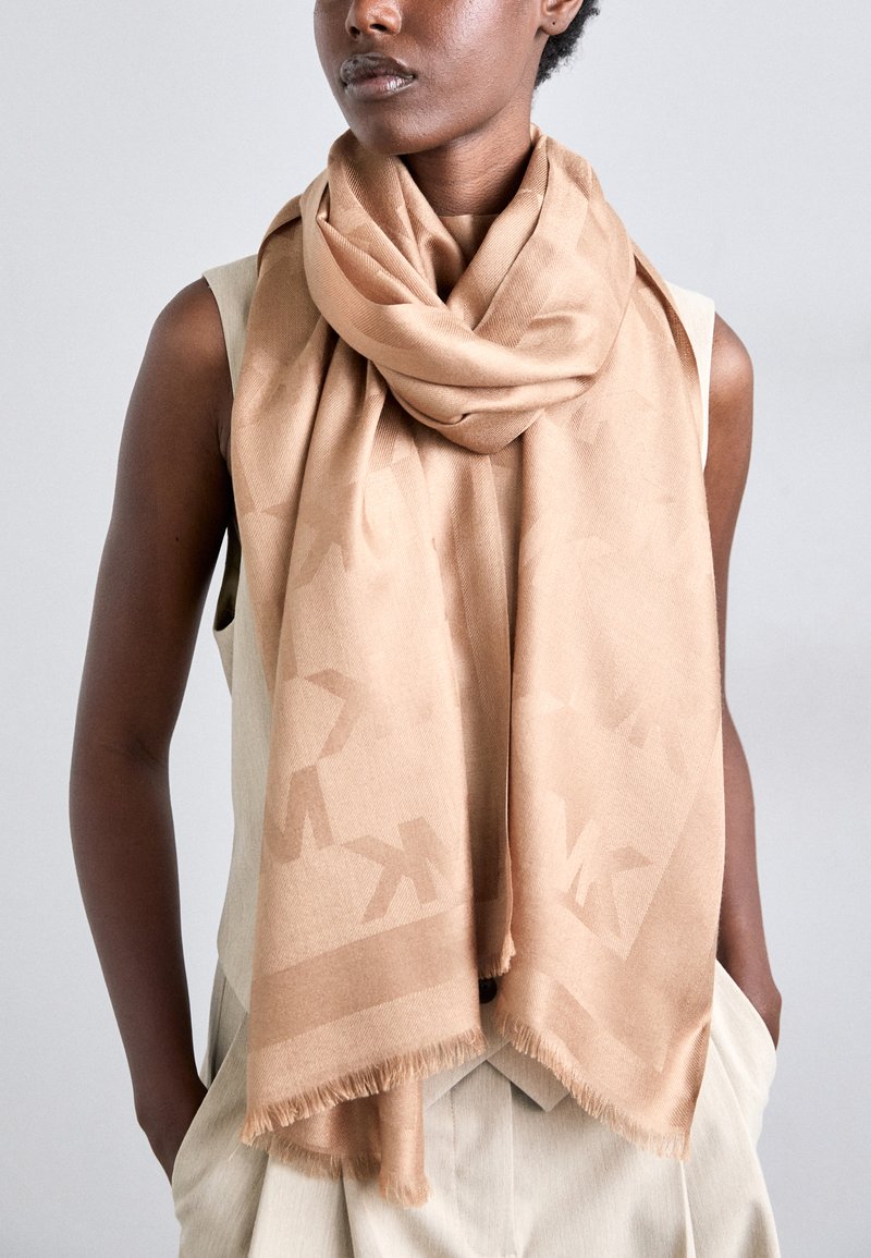 Bufanda beige hecha de tela suave, con un patrón sutil y bordes con flecos, drapeada alrededor del cuello, complementando una blusa sin mangas.