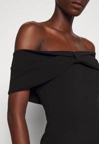 Personne à la peau foncée portant une robe noire dénudée aux épaules avec un détail de nœud sur la poitrine sur un fond clair uni.